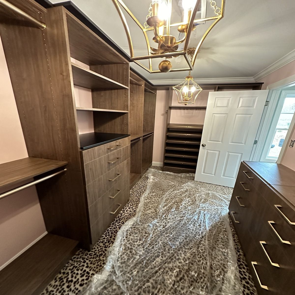 A newly built walk-in closet with wooden shelving and drawers, a patterned rug, and a chandelier. A white door is visible.