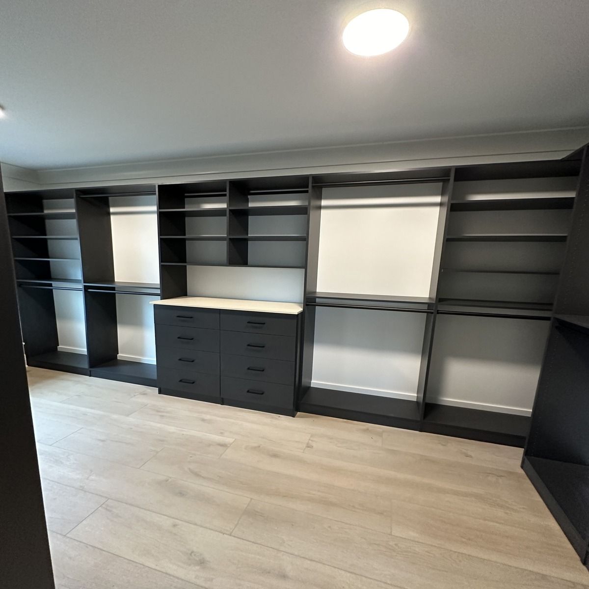 Built-in black closet with shelving, hanging rods, and a chest of drawers against white walls and light wood floors.