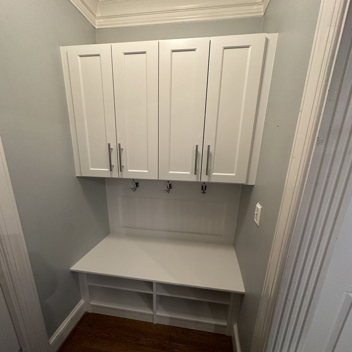 White built-in storage with upper cabinets and a bench. Hooks are mounted below the cabinets.  The walls are gray, and the floor is wood.