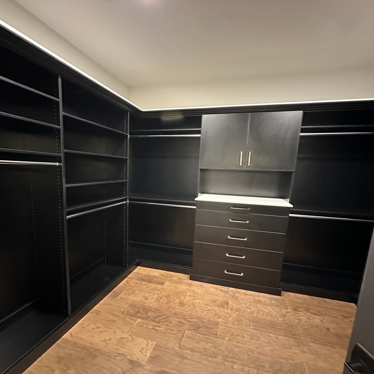 A dark, organized walk-in closet with shelves, drawers, and hanging rods. The floor is wood-toned, and the walls and cabinets are dark.