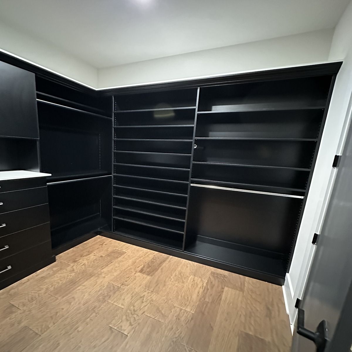 A black walk-in closet with built-in shelving and drawers. The room has light brown flooring and a white ceiling.