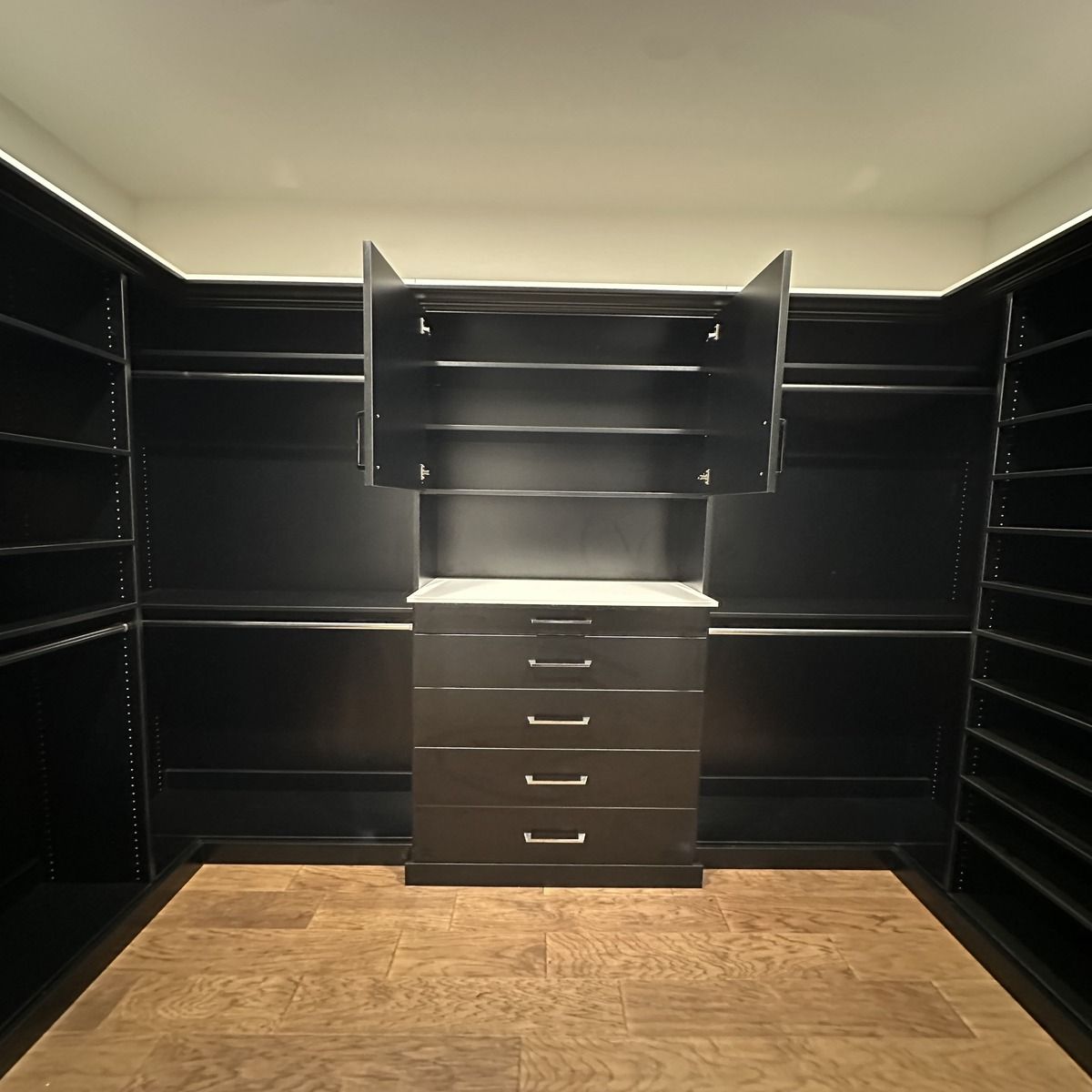 Walk-in closet with black shelving, drawers, and a central cabinet. Light-colored wood floors contrast the dark surfaces.