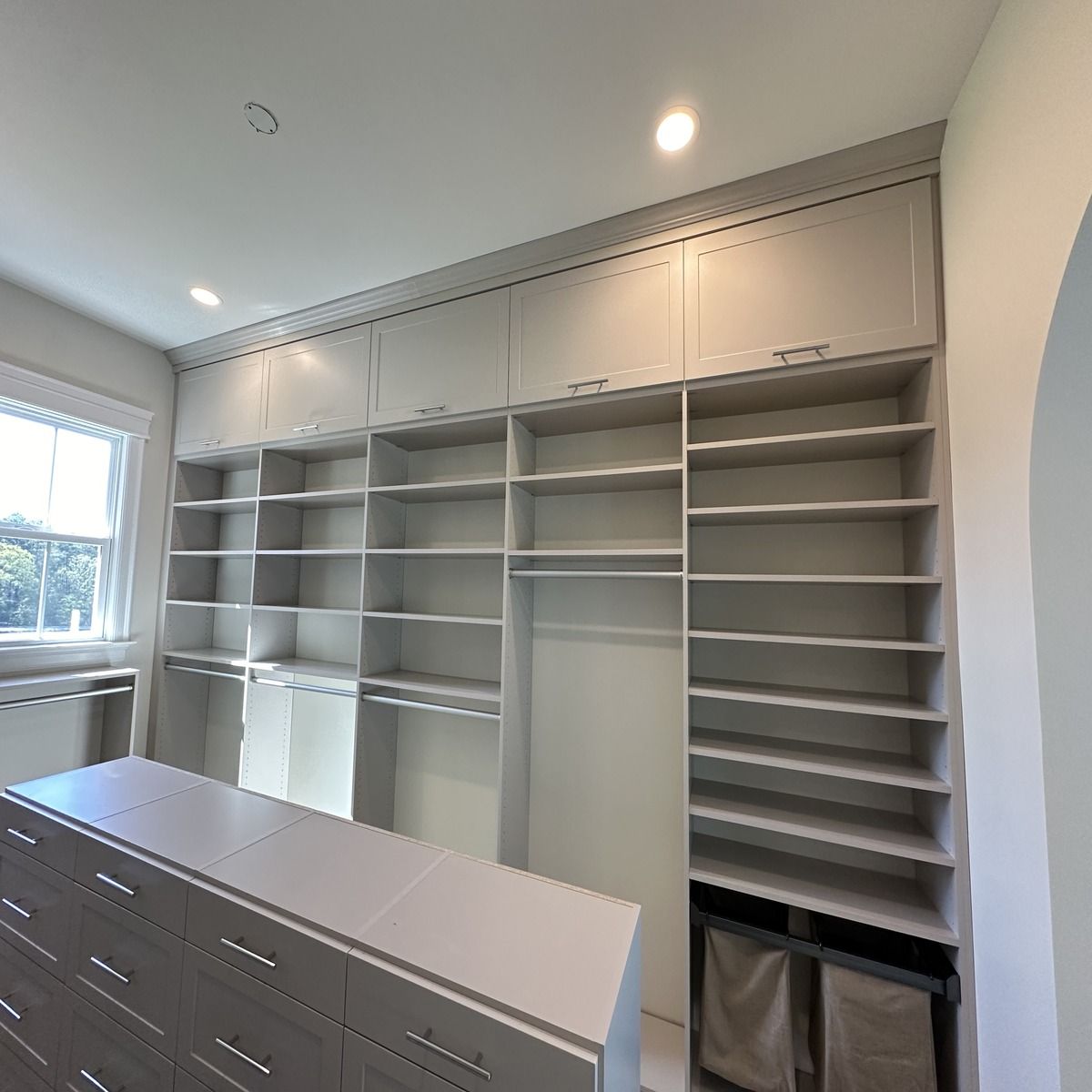 A light gray custom closet with shelving, drawers, and cabinets. A window is to the left.