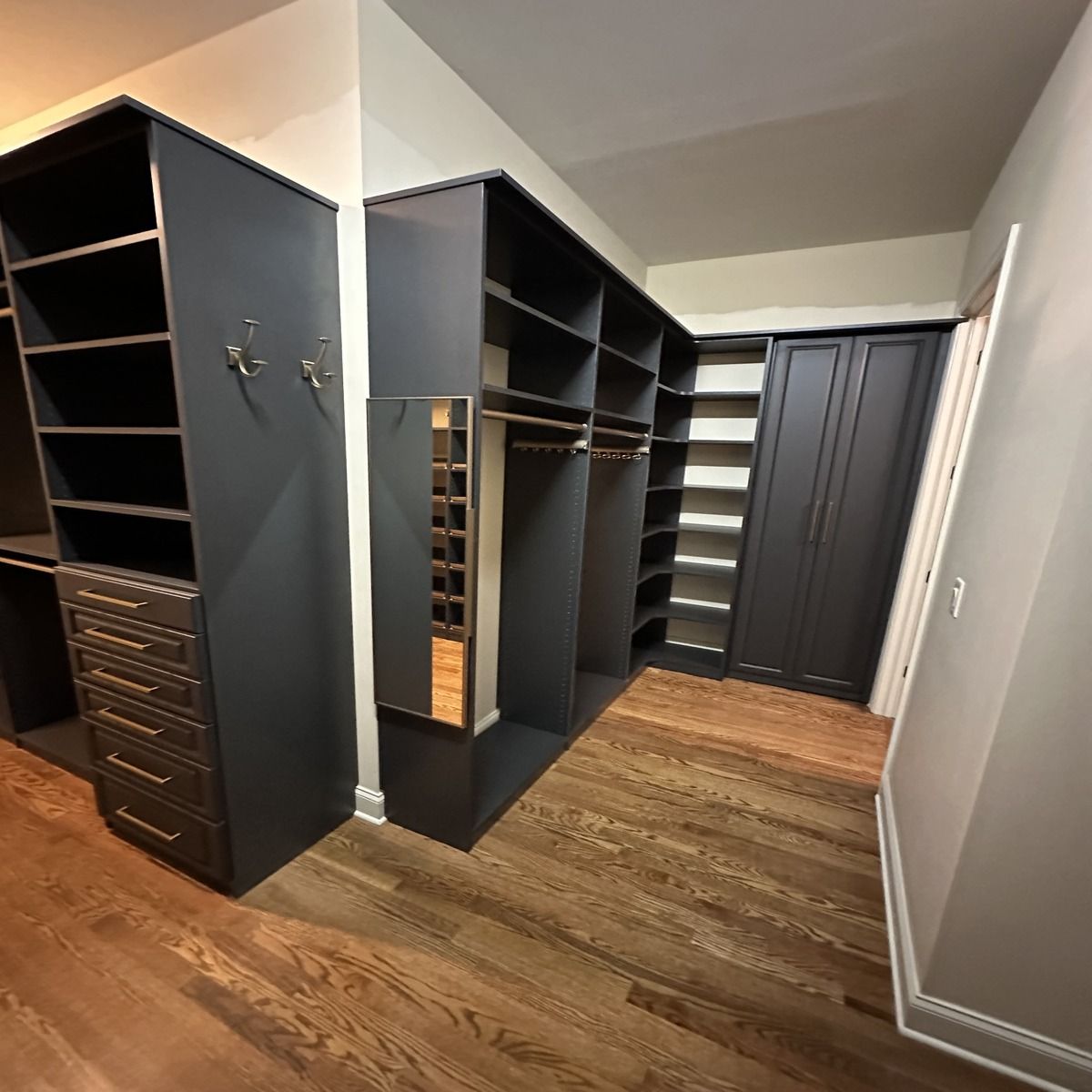 A dark custom closet with shelves, drawers, and hanging rods, featuring a full-length mirror and hardwood floors.