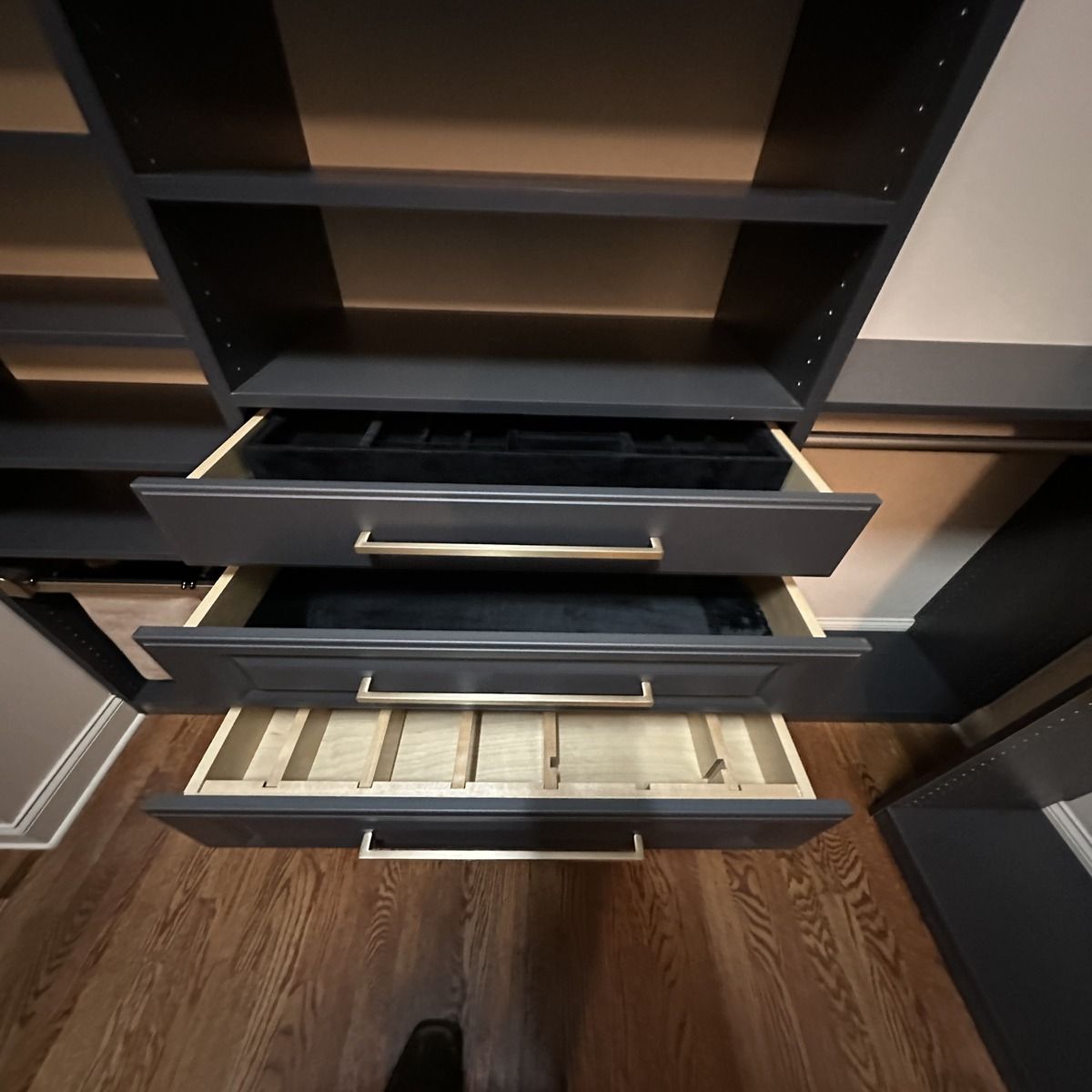 Dark gray closet drawers with gold handles, partially open, revealing jewelry organizers inside. Wooden shelves and flooring.