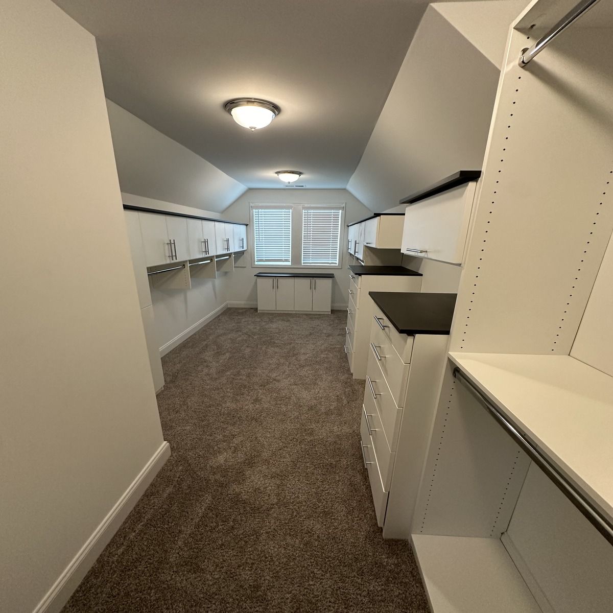 A white, spacious walk-in closet with carpet flooring, built-in shelves, cabinets, and lights. There is a window at the back.