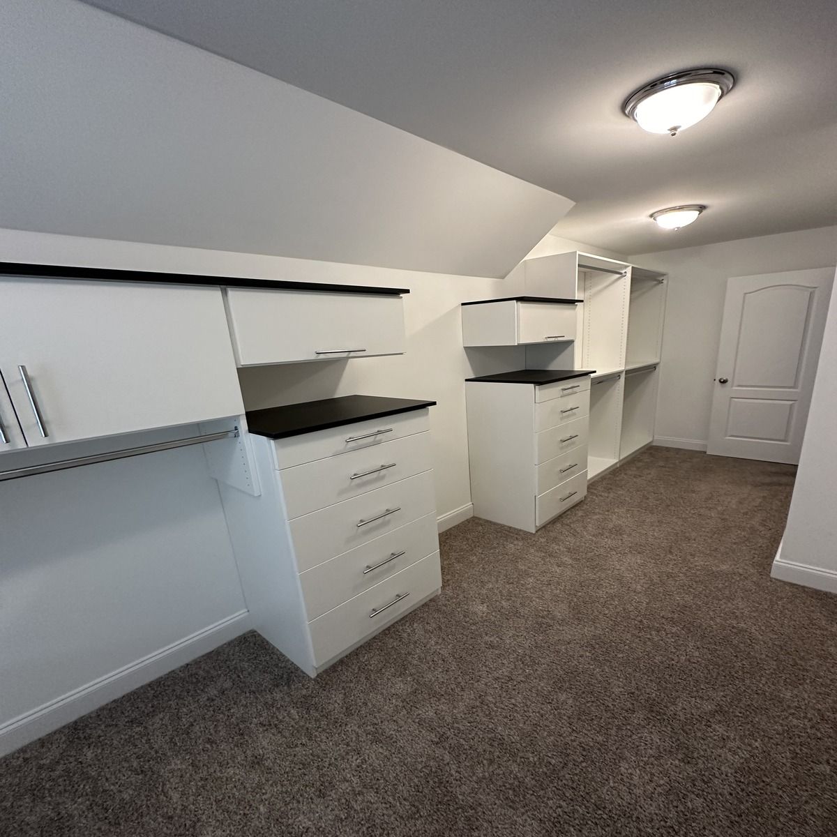 A well-organized white closet with shelves and drawers in a room with carpet. Two ceiling lights illuminate the space.