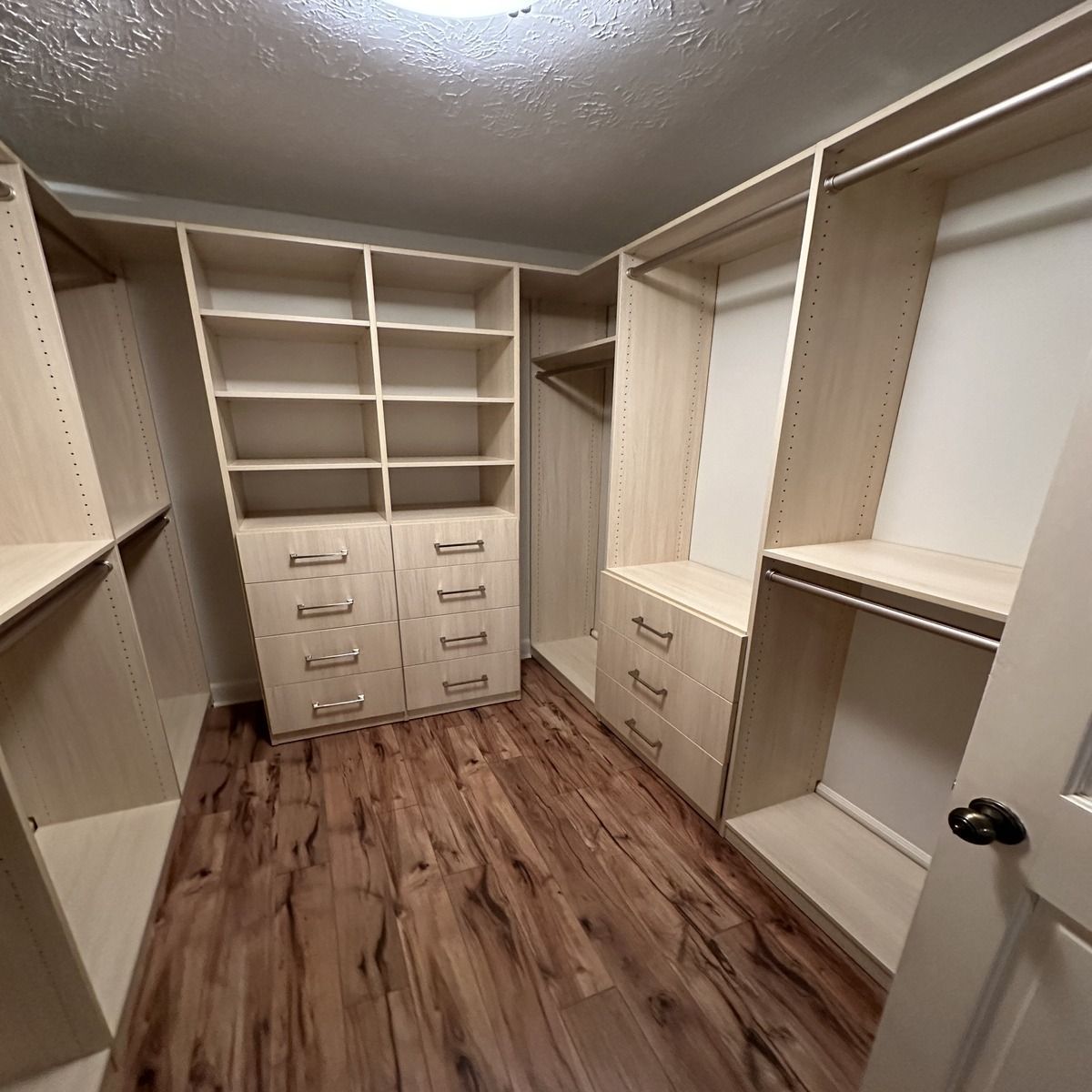 A walk-in closet with light wood shelving and drawers on a wood-look floor.