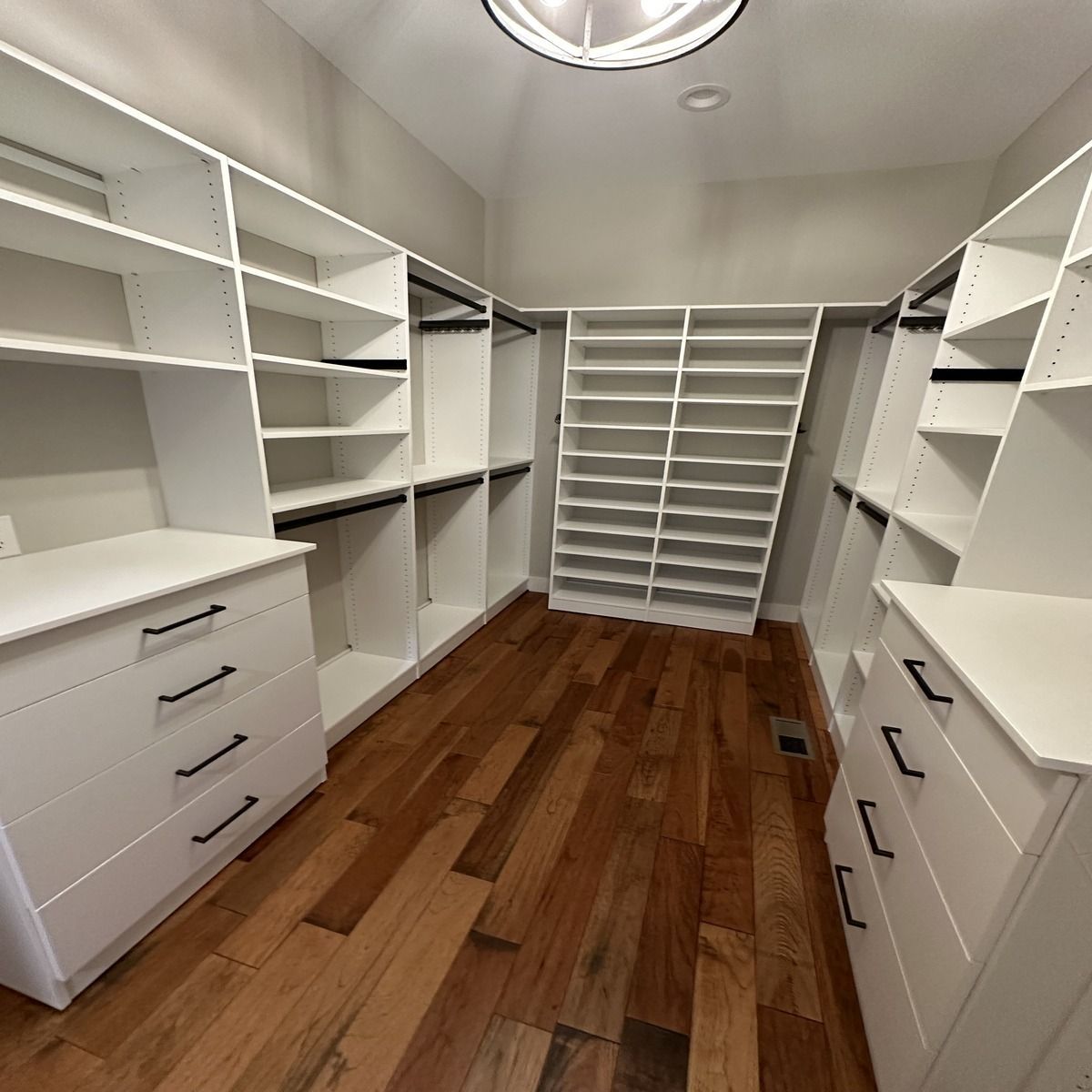 Empty white walk-in closet with built-in shelving and drawers, hardwood floors, and hanging rods.