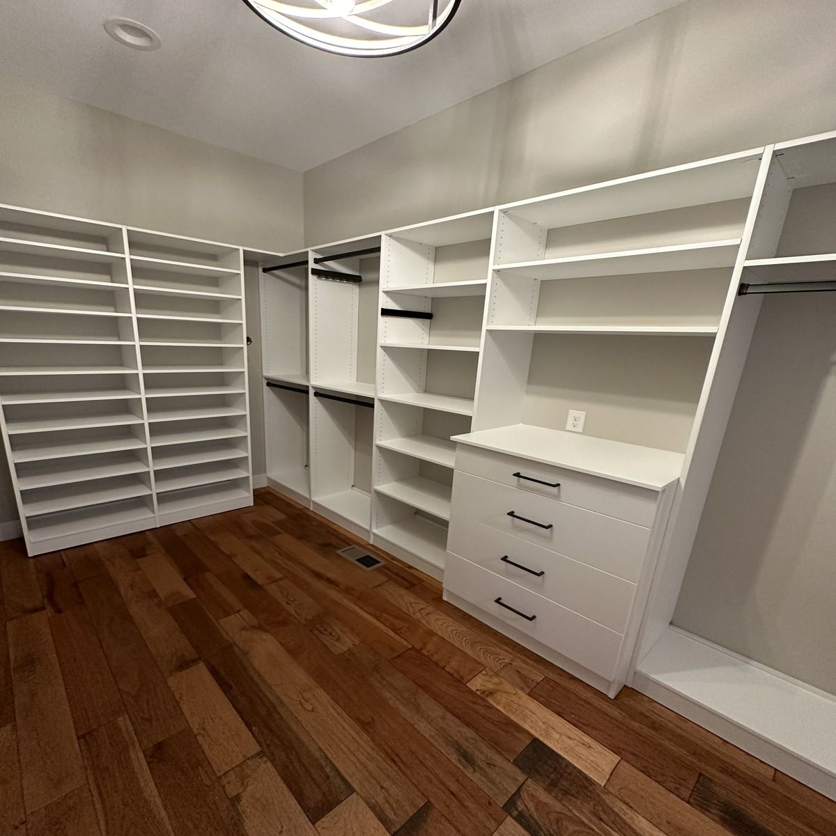 A spacious, white walk-in closet with built-in shelves, drawers, and hanging rods. The room has hardwood floors and is well-lit.
