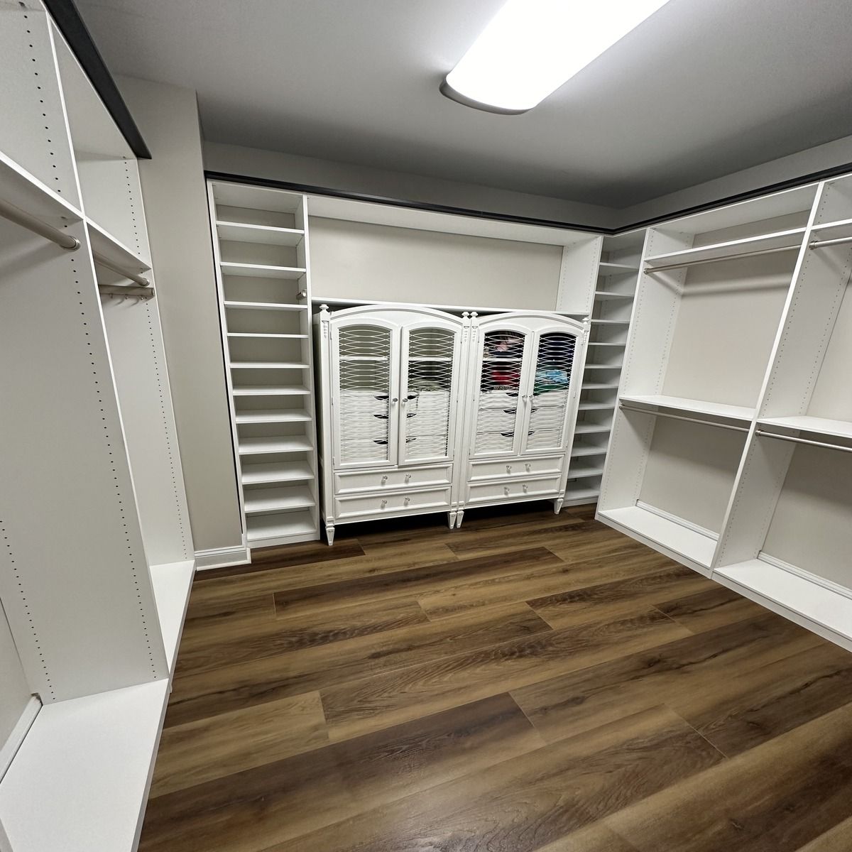 Spacious walk-in closet with white shelving and a central white cabinet against a wooden floor.