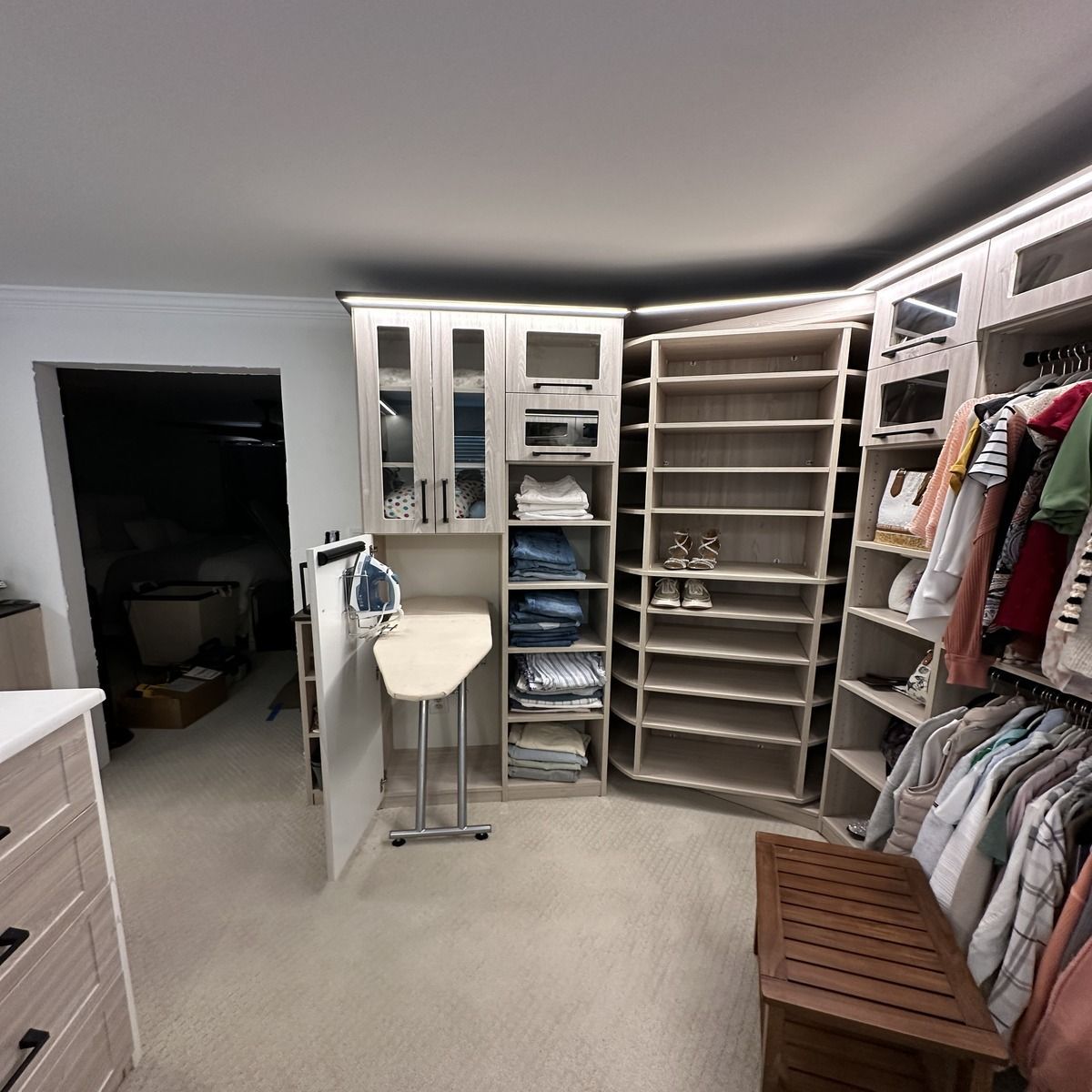A well-lit walk-in closet featuring built-in shelving, clothing racks, and an ironing board, with a wooden bench in the foreground.