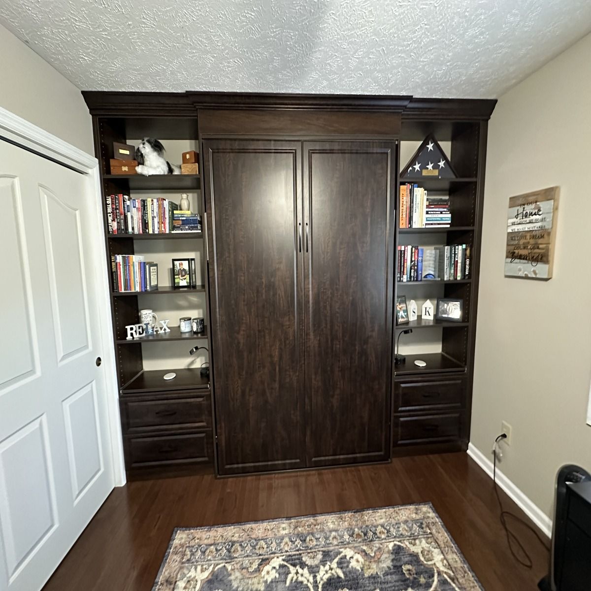 Dark wood wall unit with a built-in Murphy bed, bookcases, and drawers, in a room with a rug and closed door.