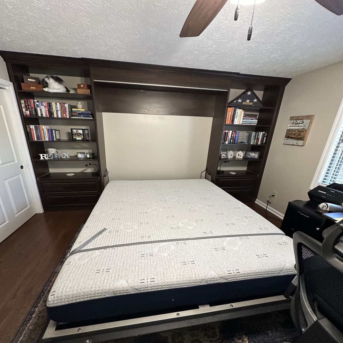 A bedroom with a pull-down bed integrated into a dark wood bookcase unit, featuring shelves, drawers, and decorative items.