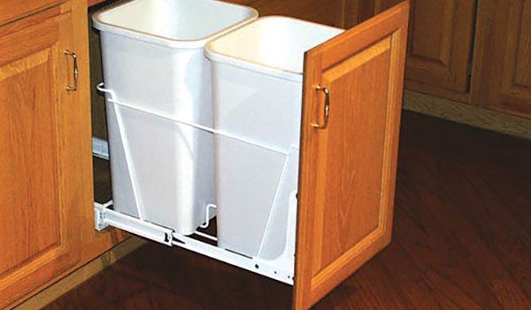 Two white trash cans are pulled out of a wooden cabinet.