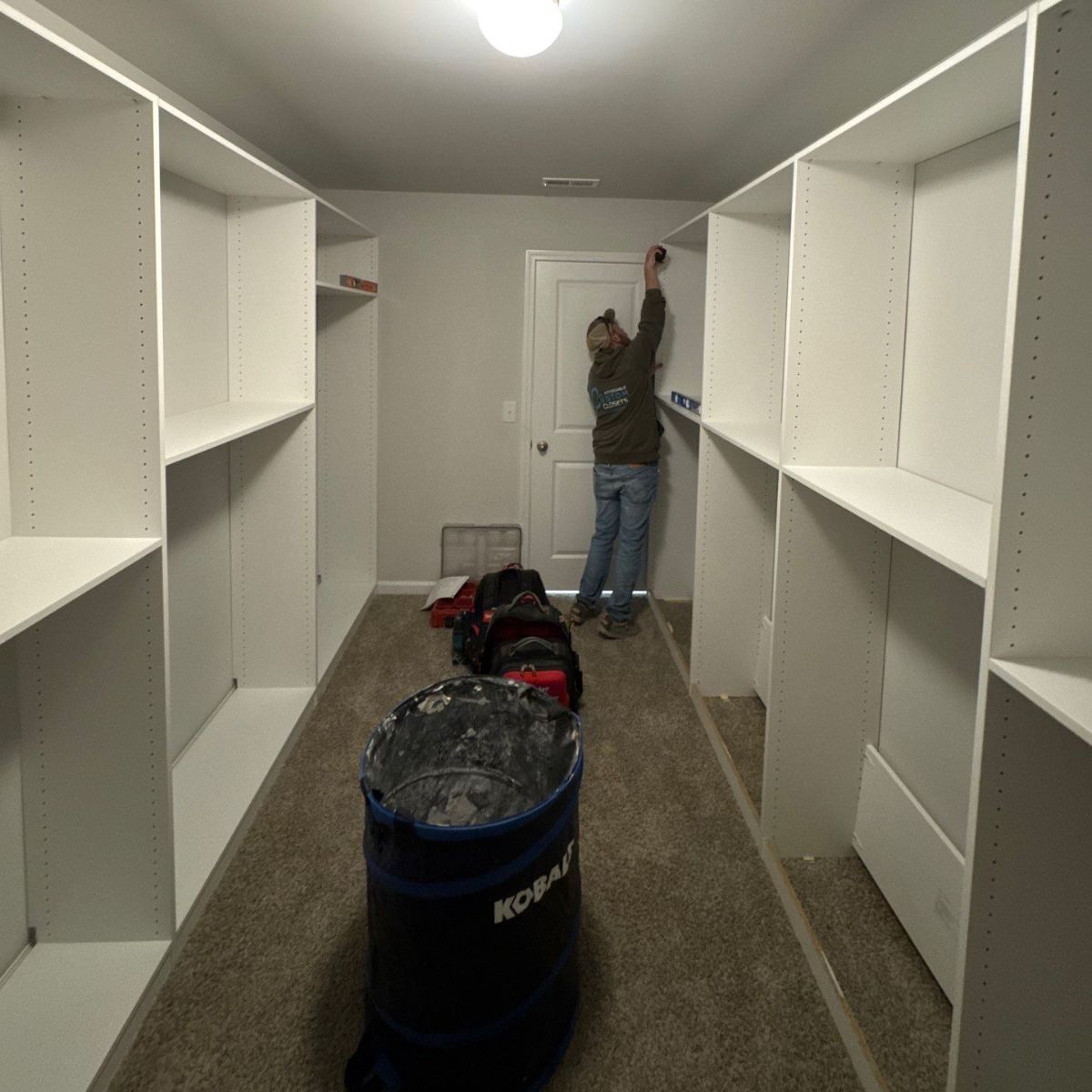 A person installing shelving in a white-walled walk-in closet. A blue trash can sits on the carpeted floor.