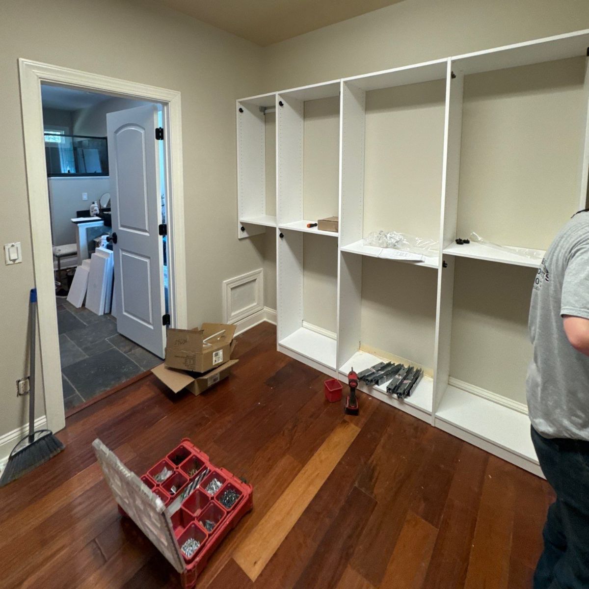 A person installs white closet shelving in a room with hardwood floors. The door to a smaller room is visible in the background.