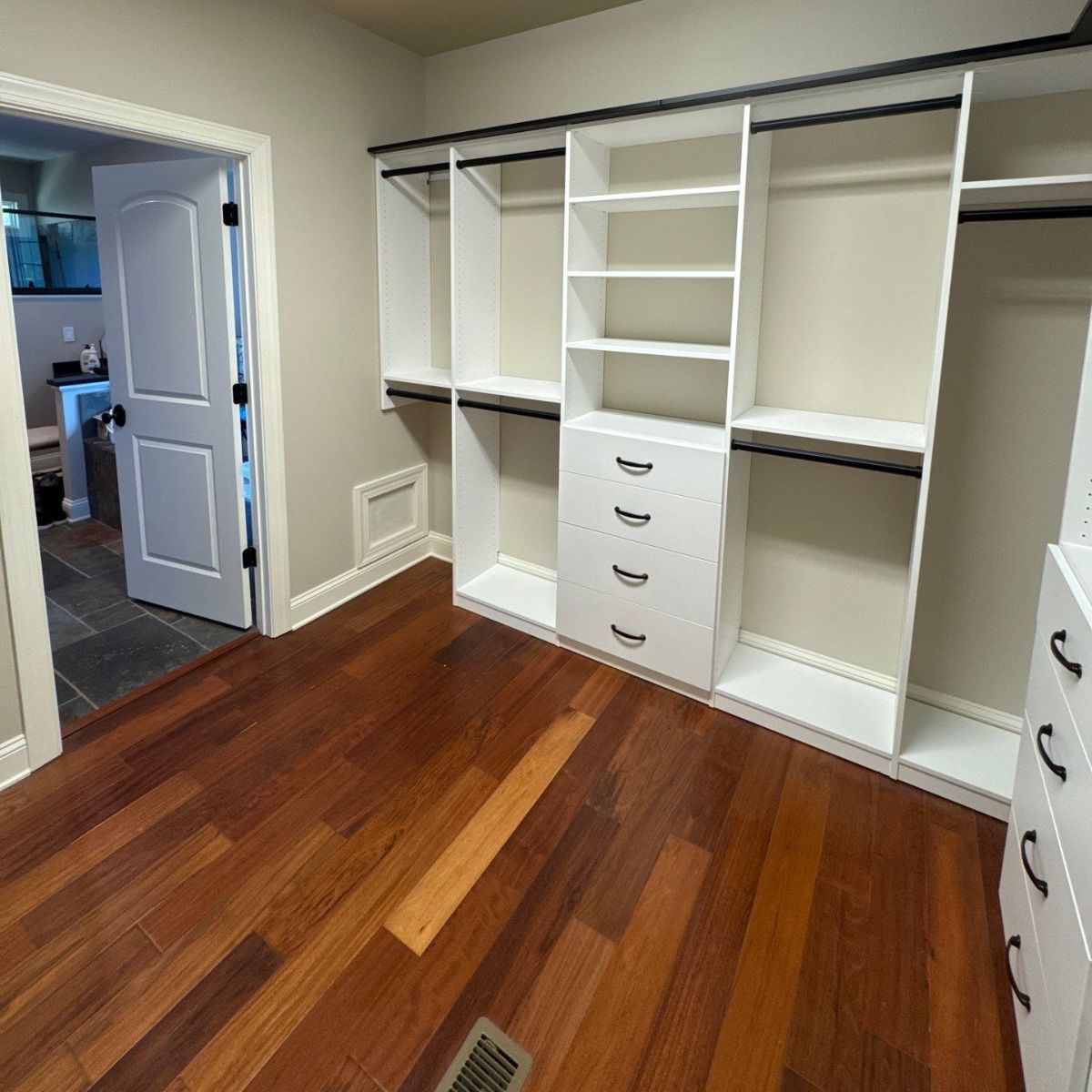 A walk-in closet with white shelving and drawers, dark hardware, and wood floors. A doorway is visible on the left.