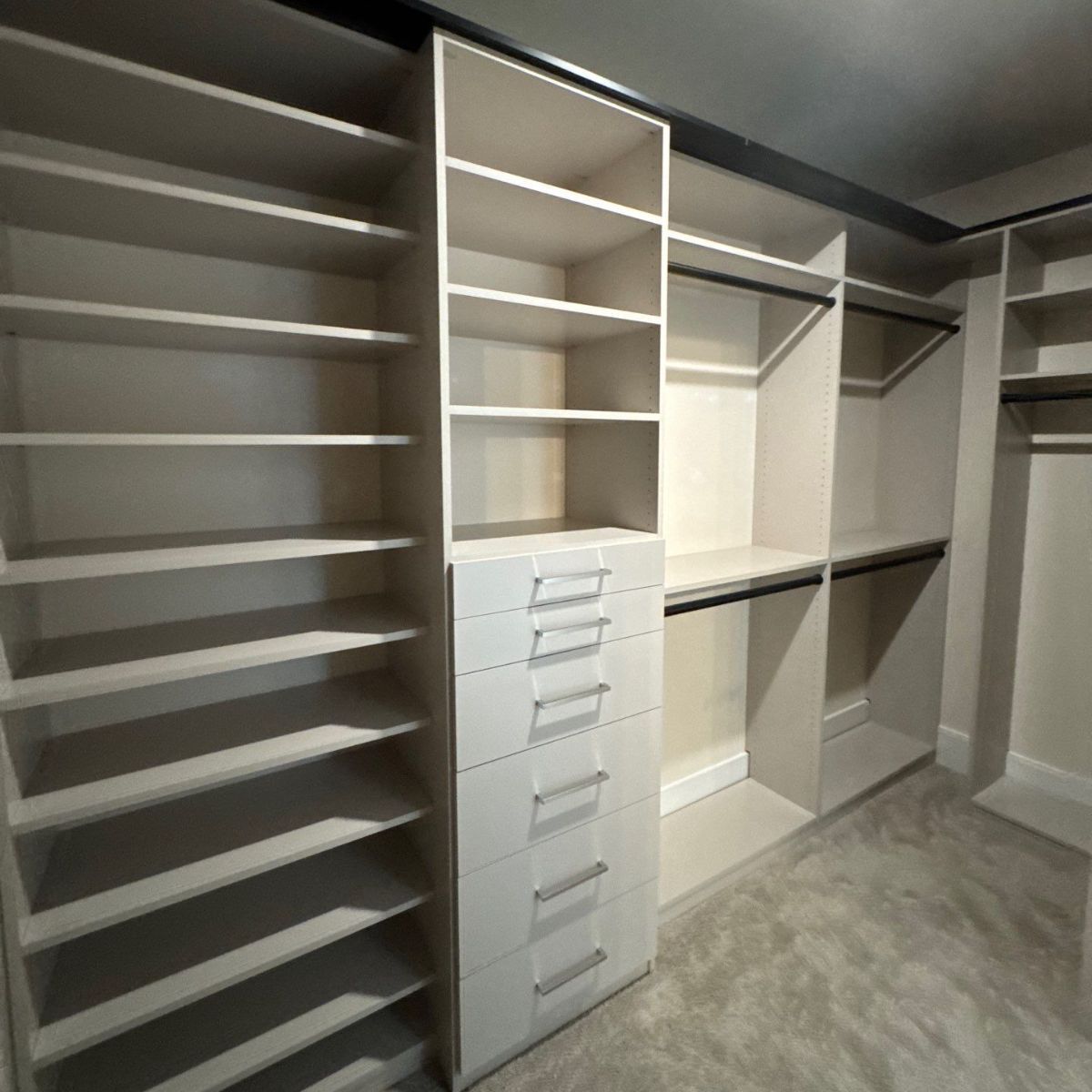 Built-in closet with white shelving, drawers, and hanging rods. The closet is empty, with a neutral carpet floor.