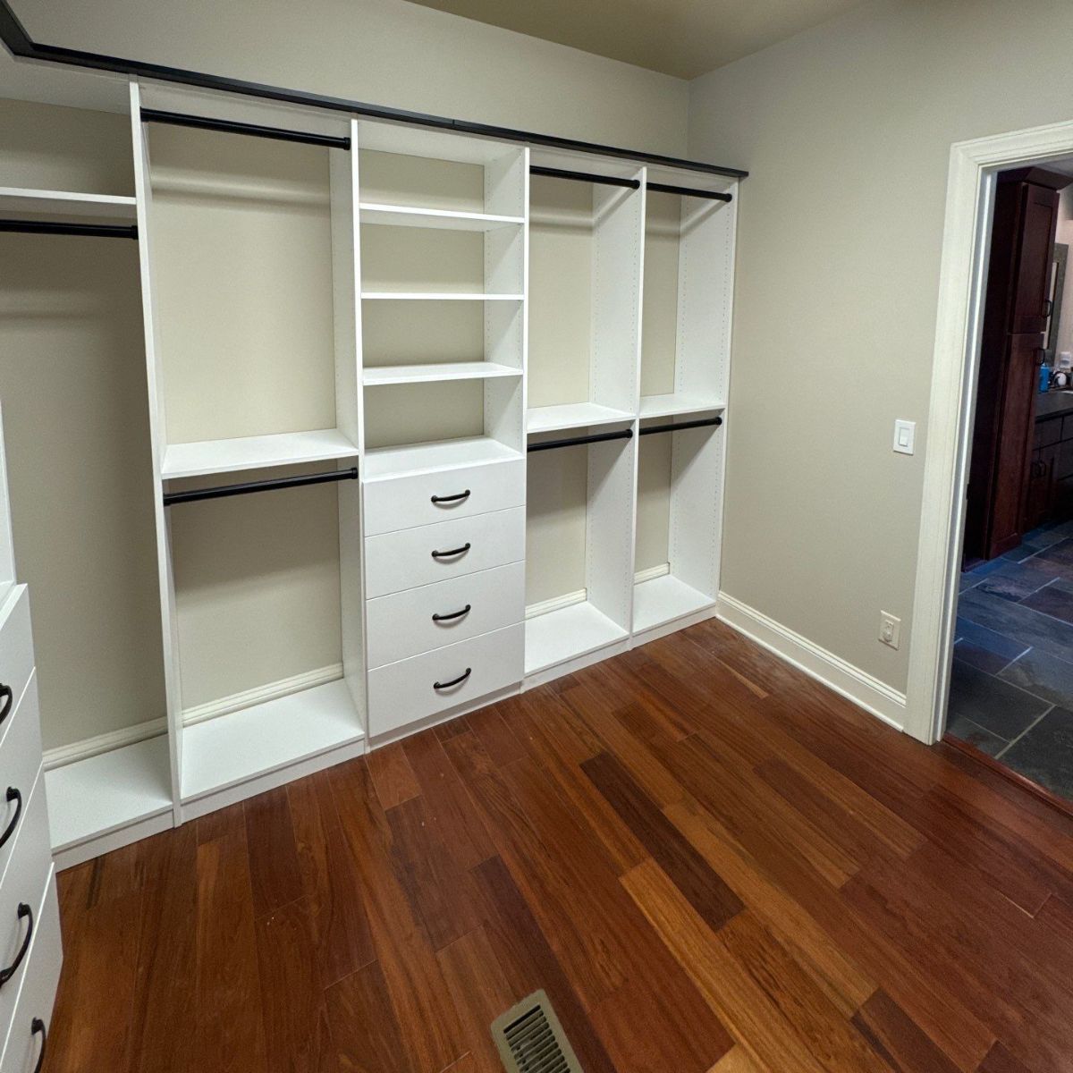 White closet organizers with black rods and pulls against a beige wall, set on a wood floor. A doorway is on the right.