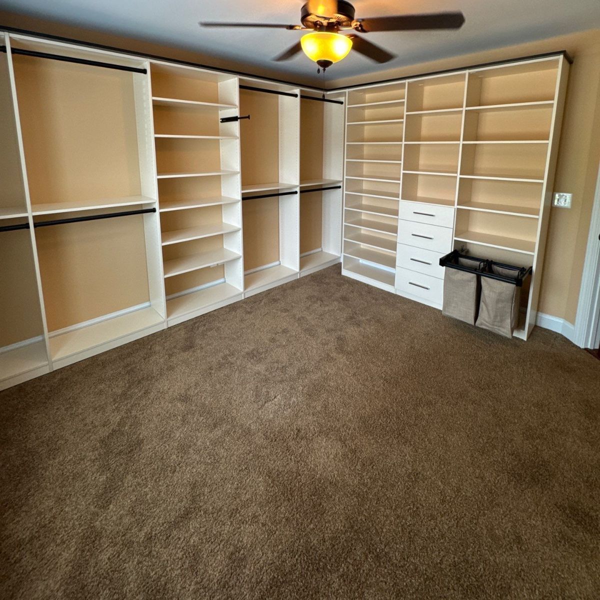 A large, empty walk-in closet with custom shelving and hanging rods. Beige walls and brown carpet.