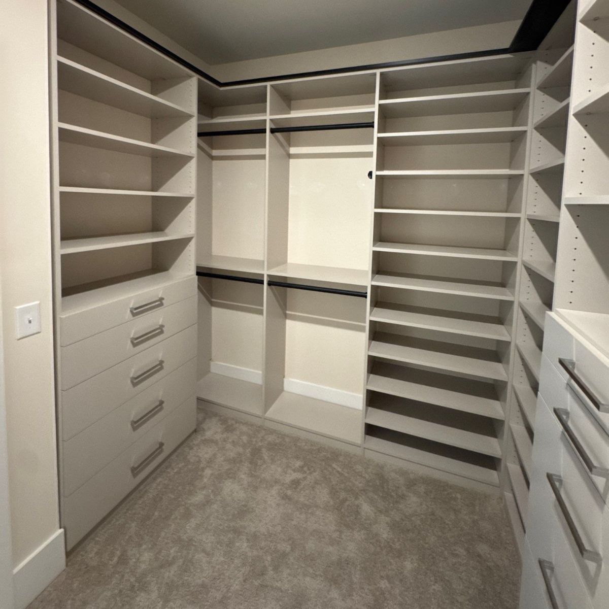 Empty walk-in closet with light-colored shelving, drawers, and hanging rods. Beige carpet covers the floor.