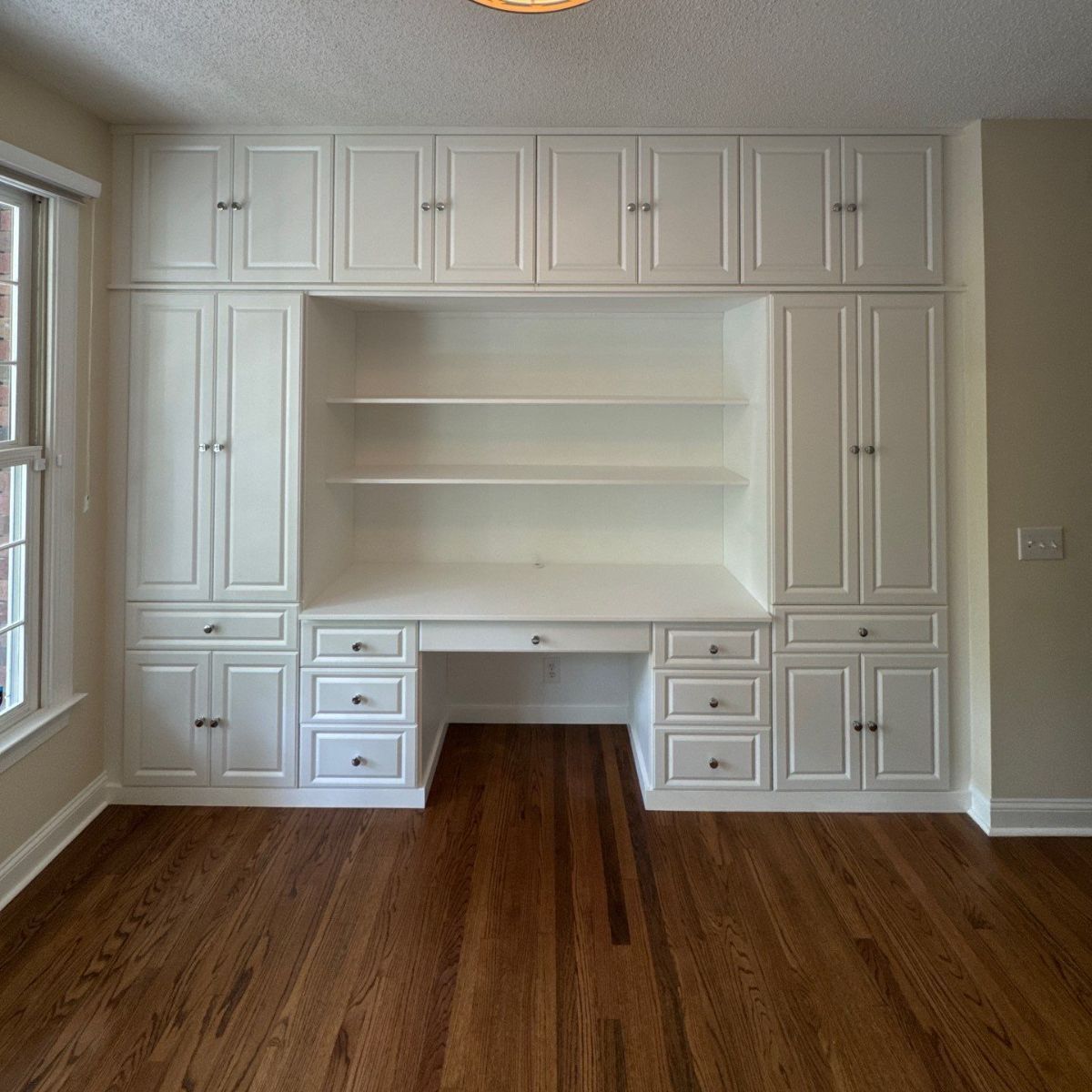 Built-in white desk and shelving unit with upper cabinets, set against a light wall in a room with hardwood floors.