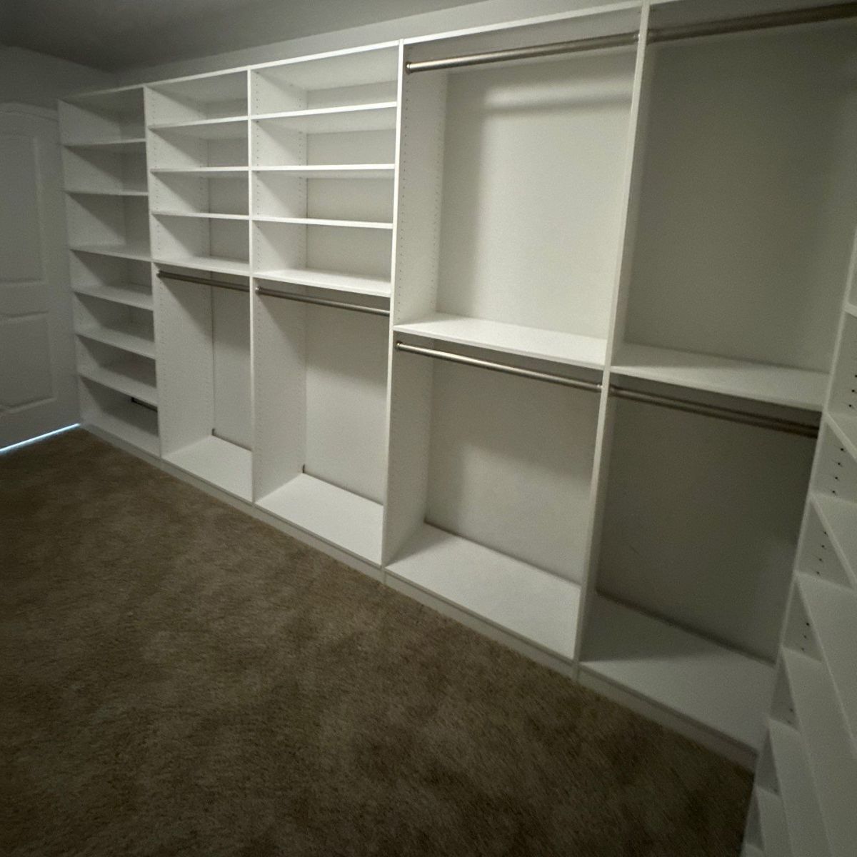 White custom closet shelving with shelves, hanging rods, and a carpeted floor. The closet is built-in, and the door is visible on the left.