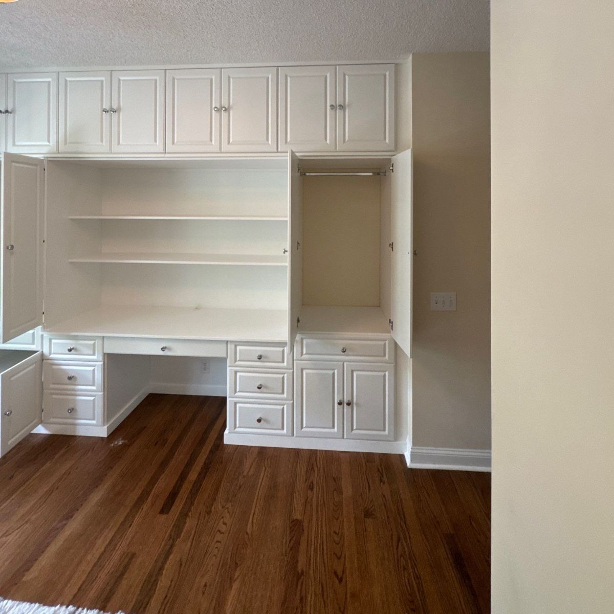 White built-in desk and storage unit with open cabinets and drawers against a pale wall; hardwood floors.
