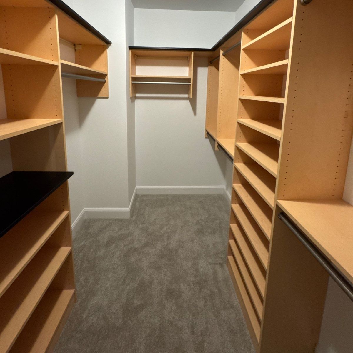 Empty walk-in closet with beige shelving and a gray carpet floor. One side has shelves and a rod, the other side has shelves.