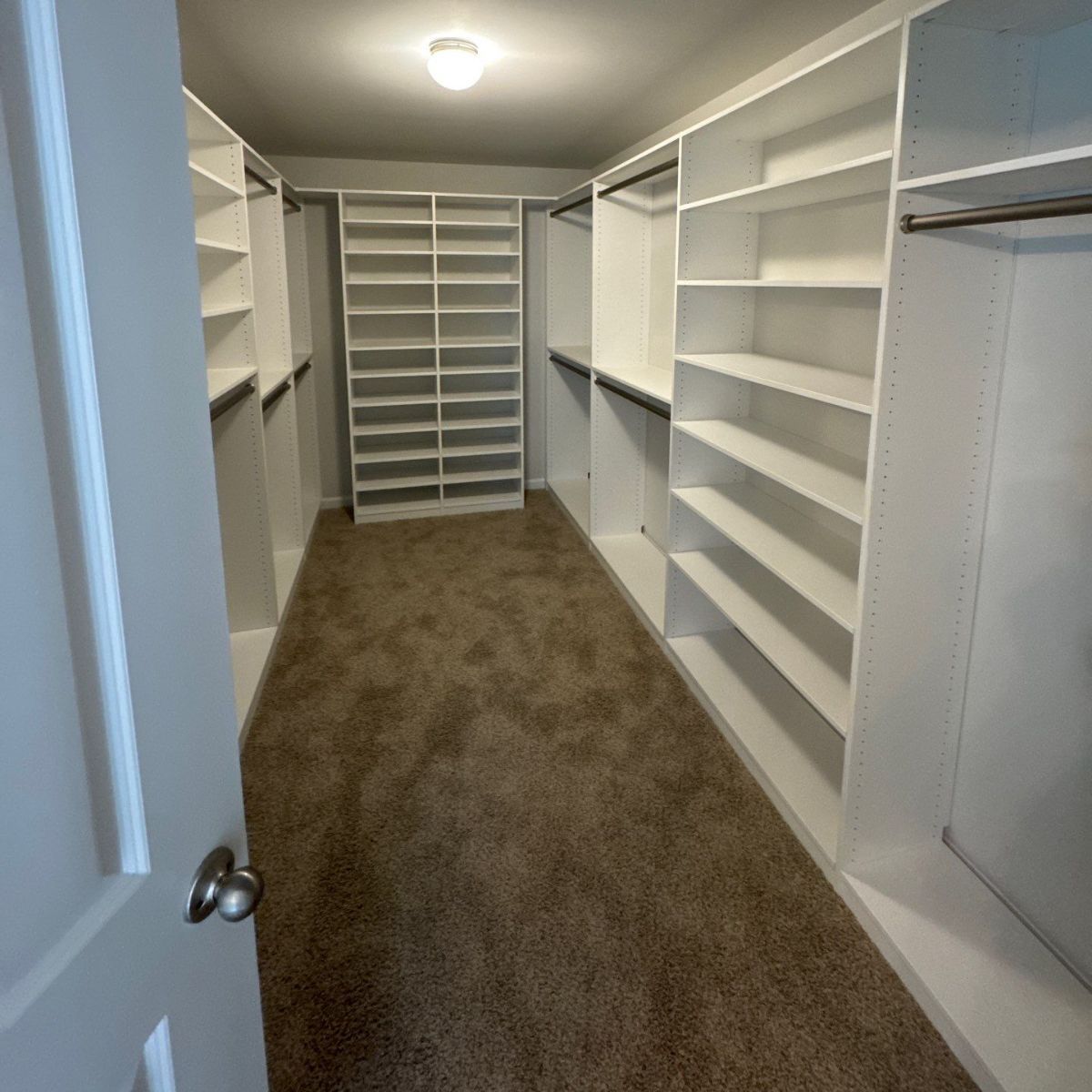 Walk-in closet with white shelving and a brown carpet. The closet has multiple shelves and hanging rods for clothing.