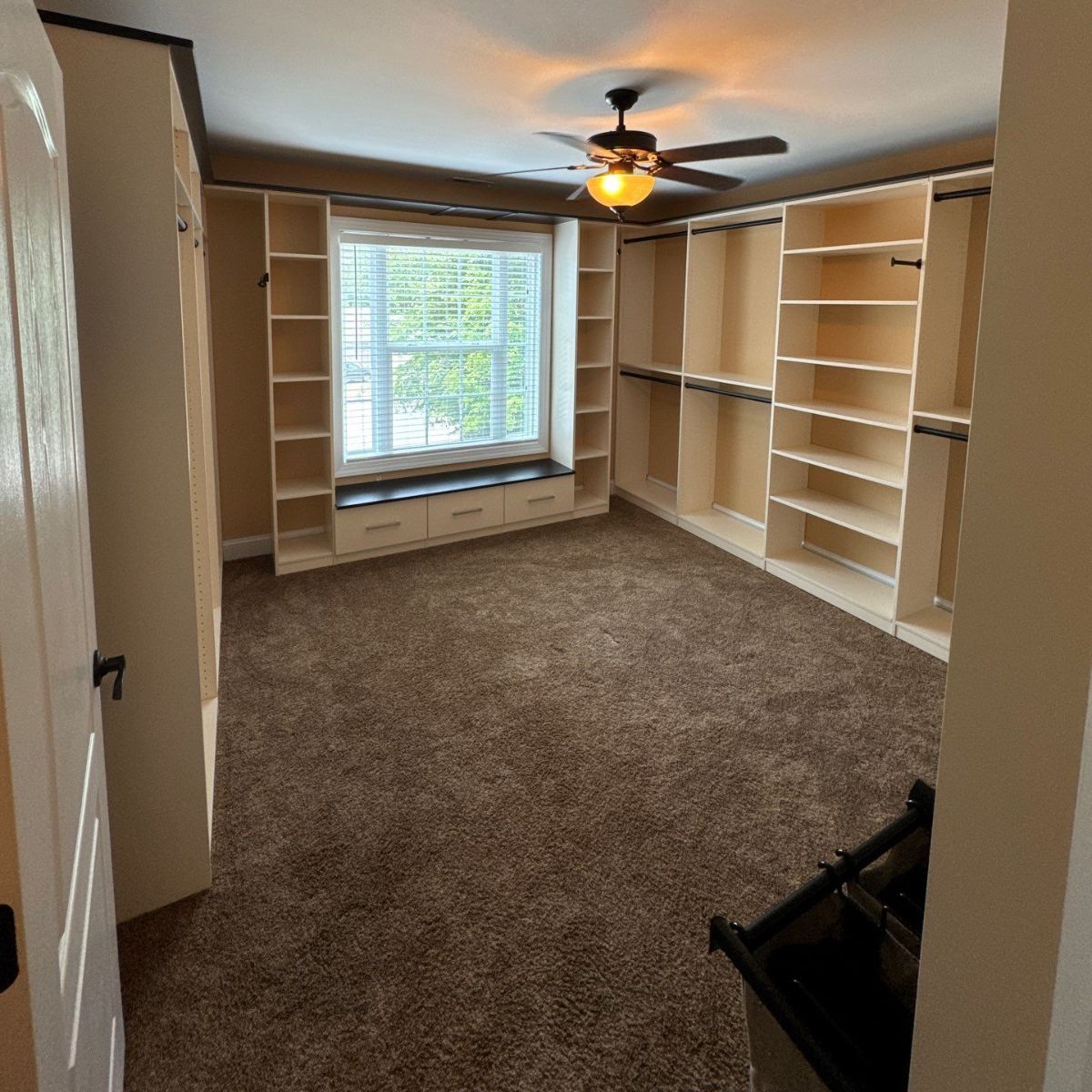 A large walk-in closet with built-in shelving, a window seat, and a ceiling fan. Brown carpet covers the floor.