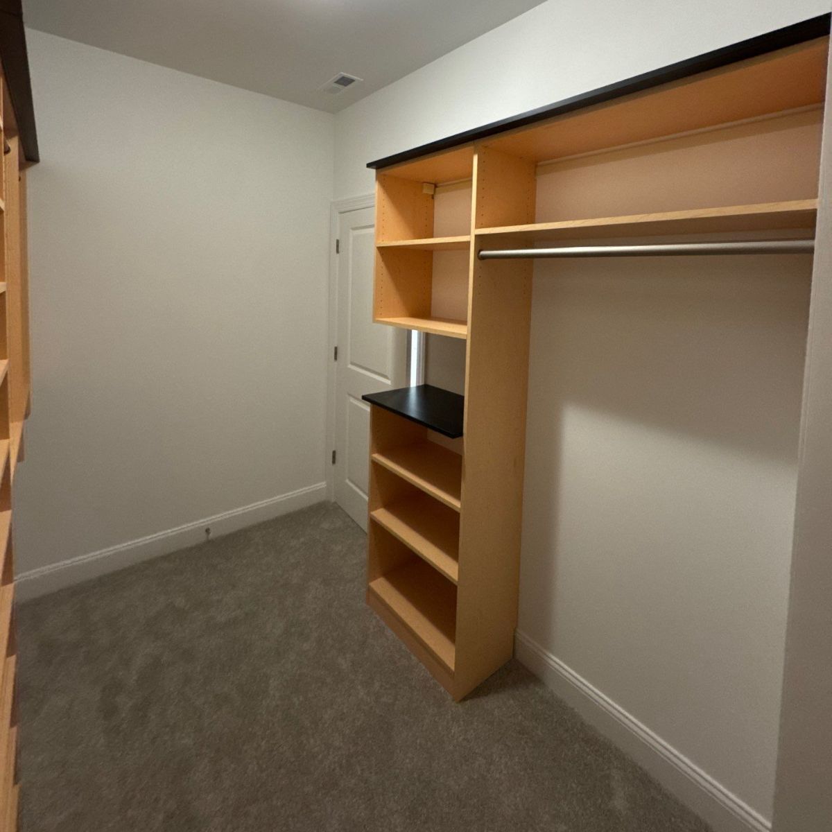 A walk-in closet with light-colored wood shelving and a hanging rod. The room has light gray carpet and white walls.