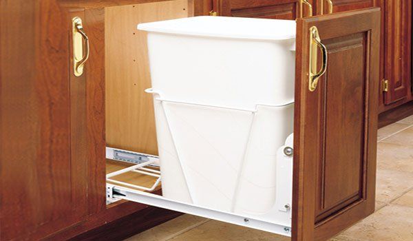 A white trash can is pulled out of a wooden cabinet.