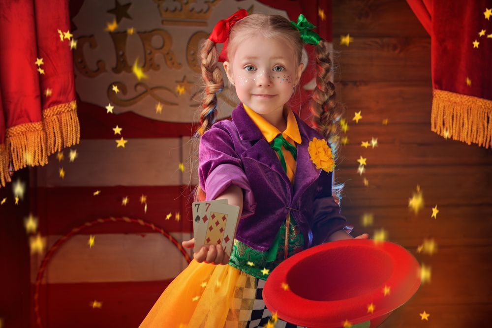 A little girl in a circus costume is holding a hat and playing cards.