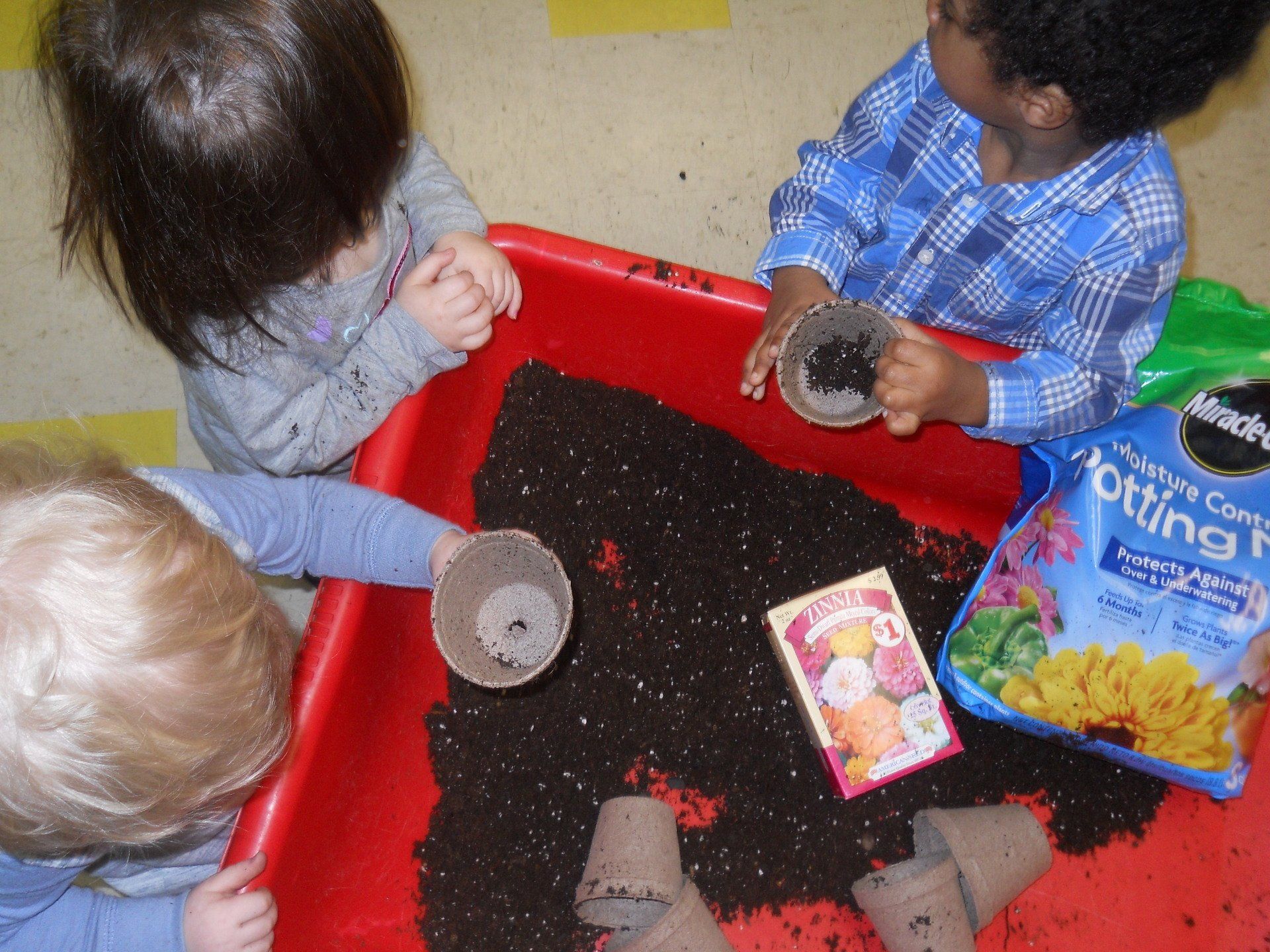 Three children are playing with potting soil in a red tray