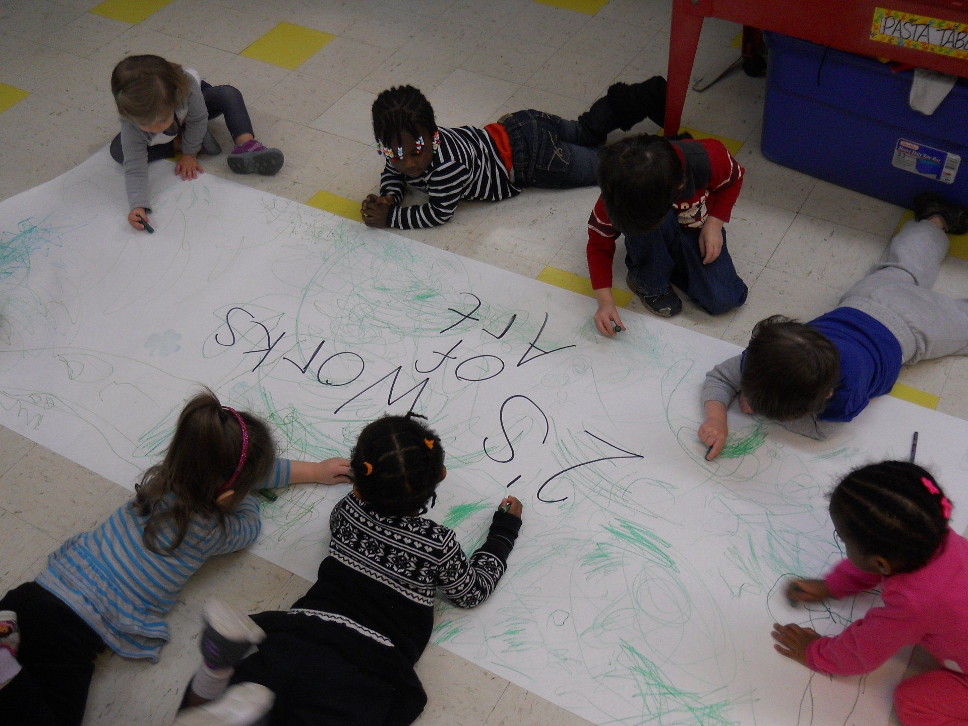 A group of children are laying on the floor writing on a large piece of paper
