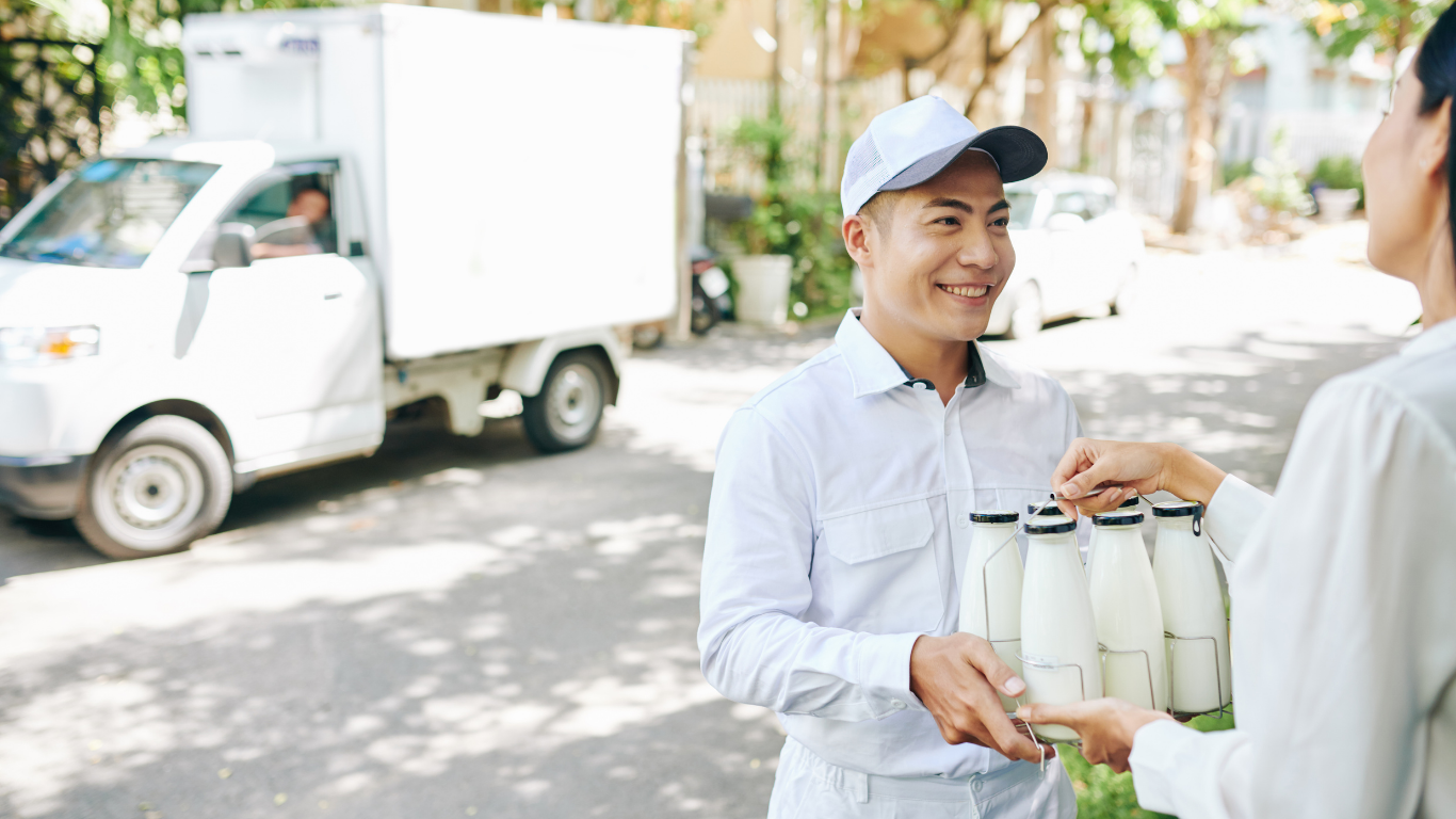 Delivery driver handing a tray of milk bottles to a person in a residential street. White delivery truck in the background.