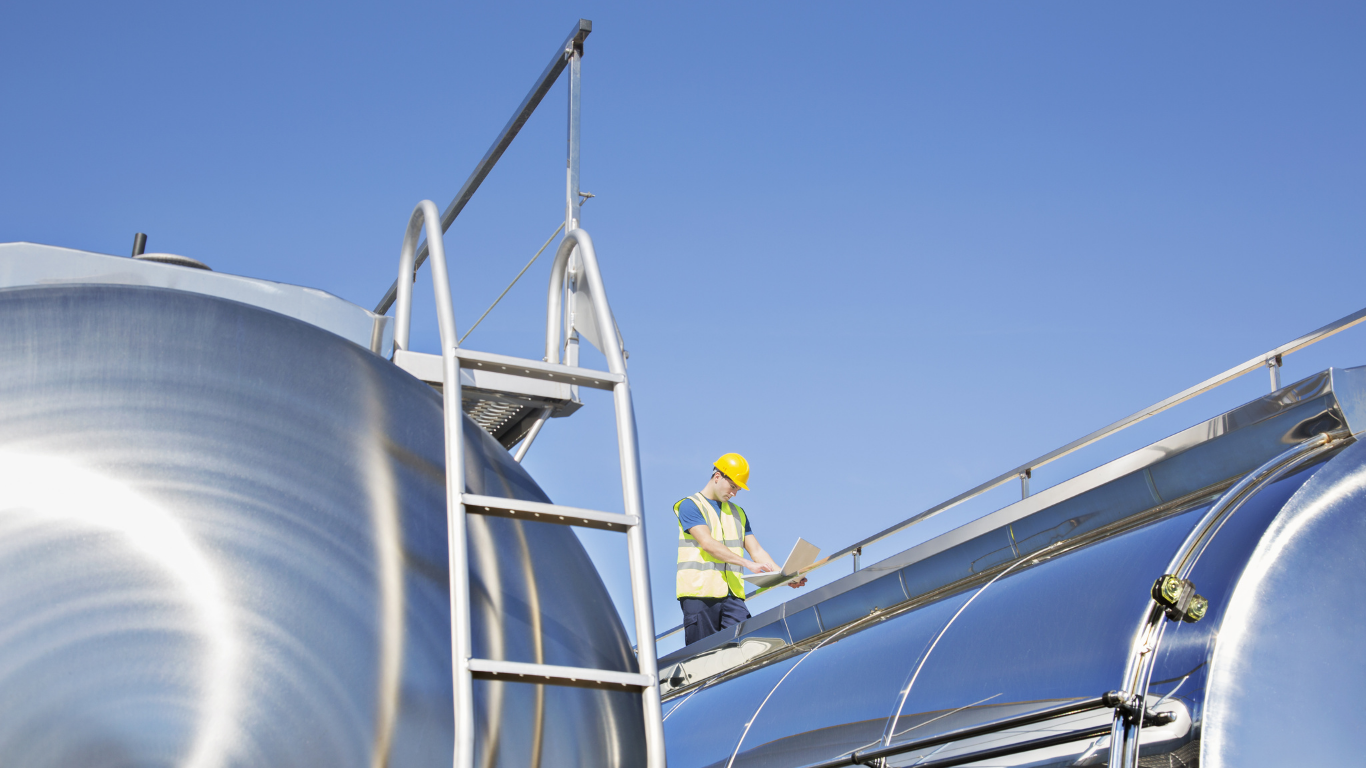 Worker in safety gear on top of a metal tanker inspecting something, blue sky background.