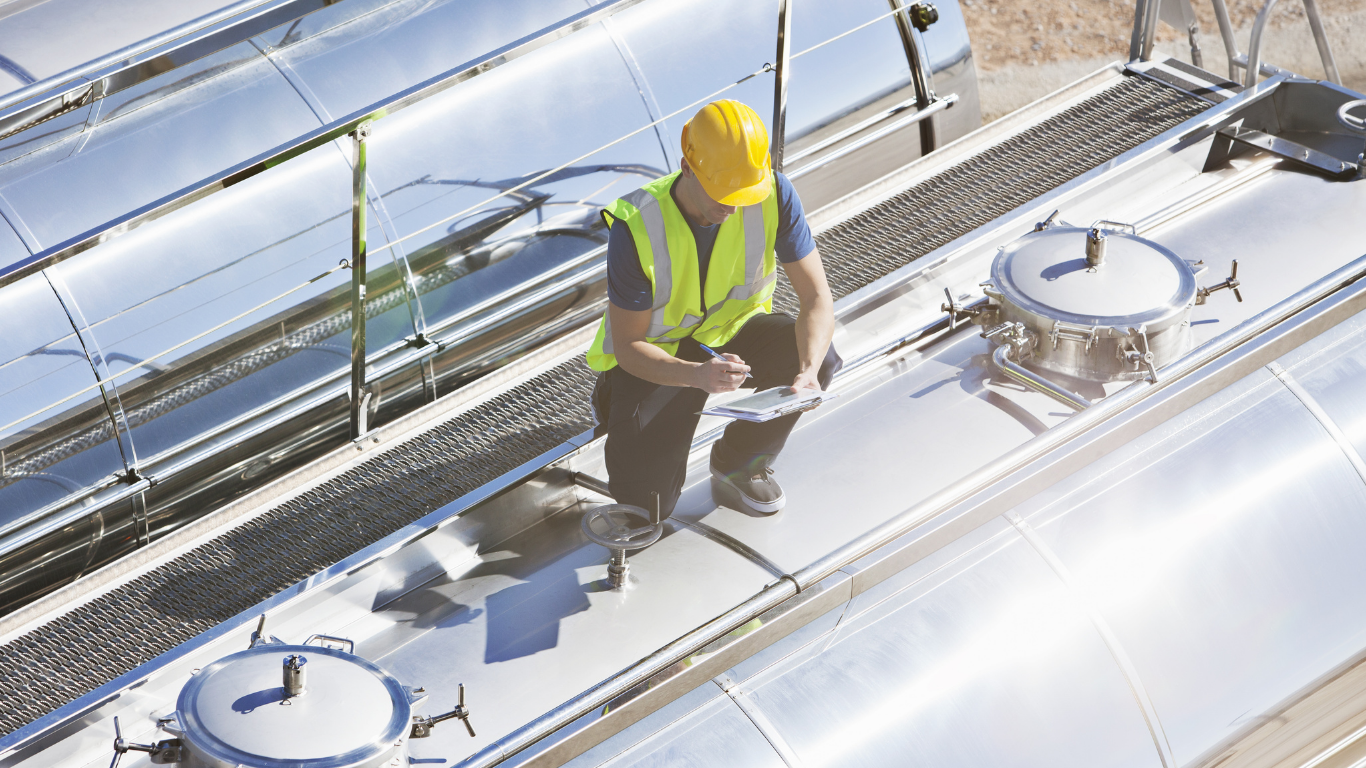 Person in yellow hard hat and vest inspects tanks on top of a tanker truck, holding a clipboard.