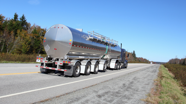 Silver tanker truck driving on a two-lane road under a blue sky, autumn trees in the background.
