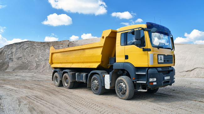 Yellow dump truck on a sandy construction site under a blue sky with clouds.