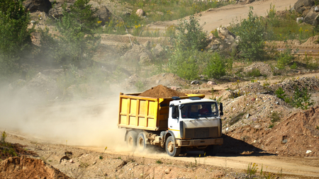 Yellow dump truck driving on dirt road, kicking up dust in a quarry.