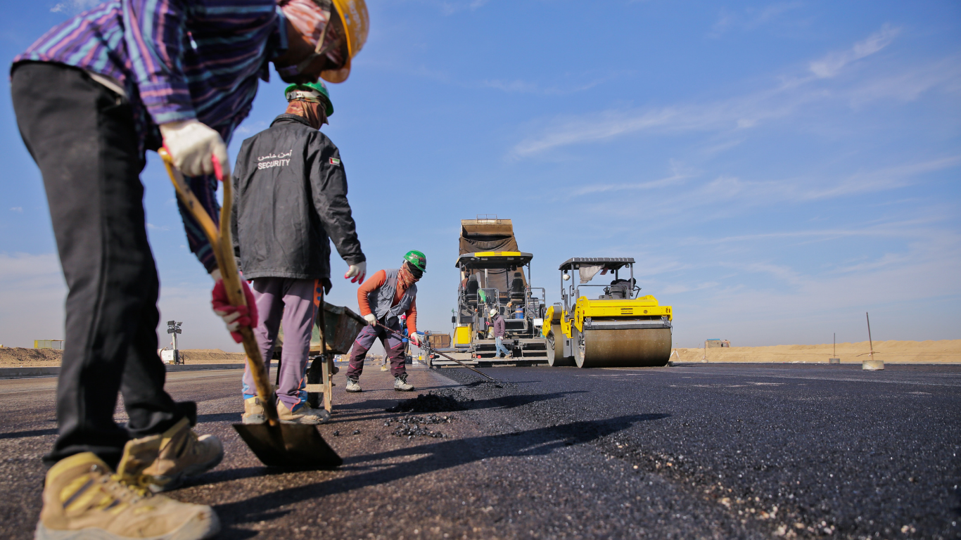 Road construction: Workers laying asphalt, with paving equipment in the background under a blue sky.
