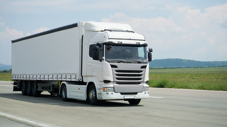 White semi-truck driving on a road, with a blue sky and green field in the background.