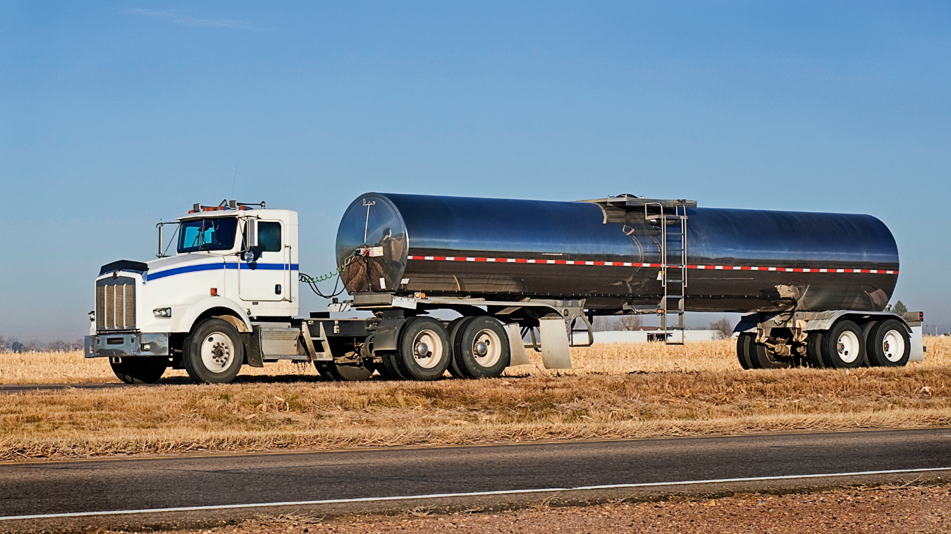 White semi-truck with a tanker trailer driving on a road under a blue sky, beside a field.
