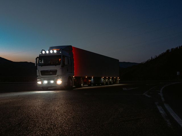 Truck on road at night, headlights on, with trailer. Dark sky and trees in background.