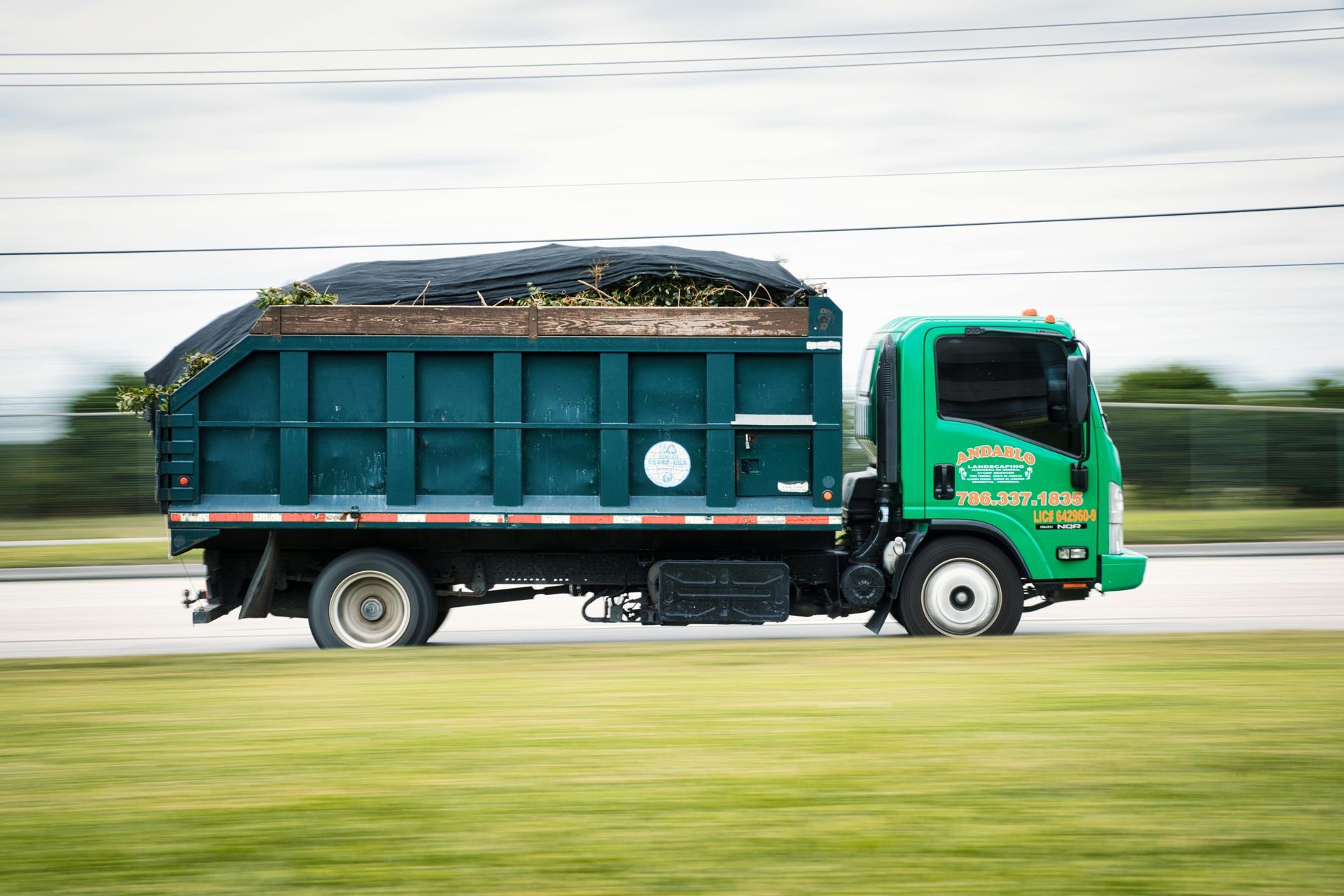 Green garbage truck driving on road, carrying yard waste.