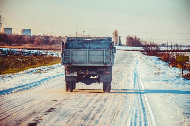 Truck driving on a snowy, rural road. Sunlit scene with a light blue sky and brown fields.