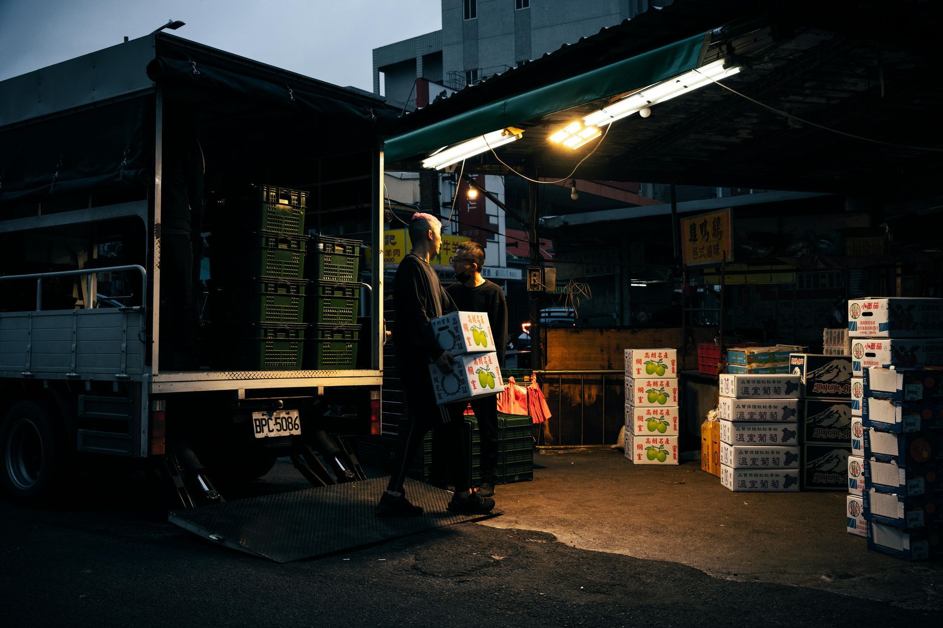 Two people unload produce from a truck at a market stall, illuminated by a light.